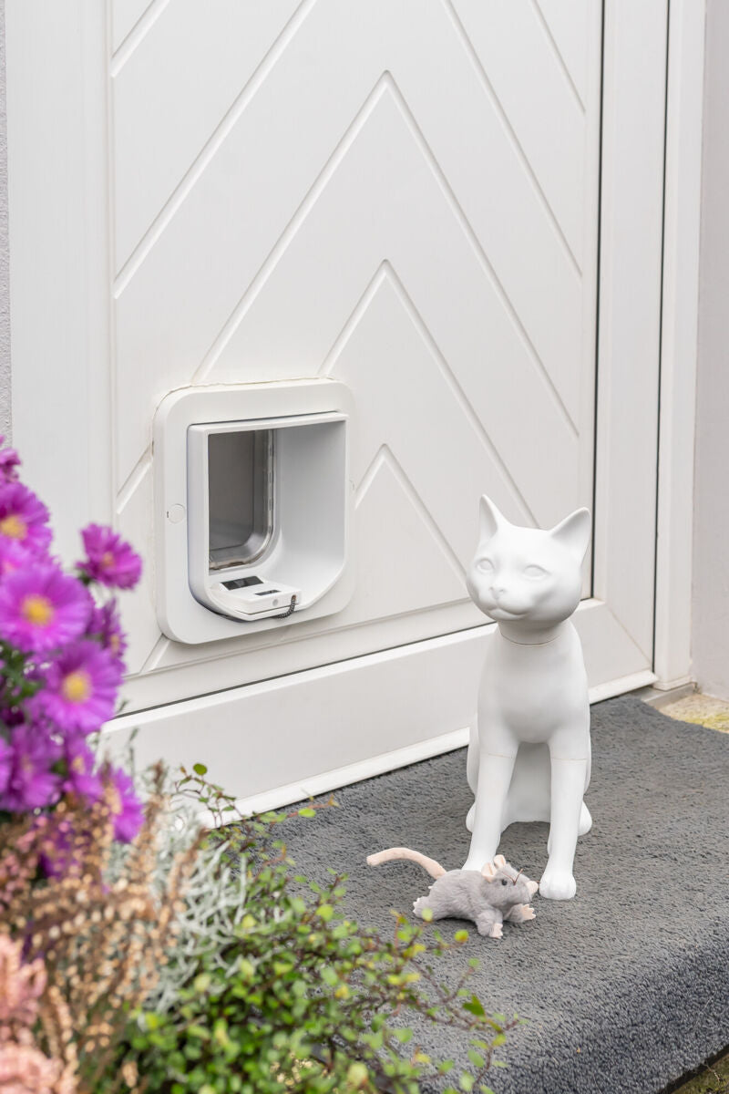 Weiße Katzenskulptur neben Katzenklappe auf grauem Teppich vor einer Tür.