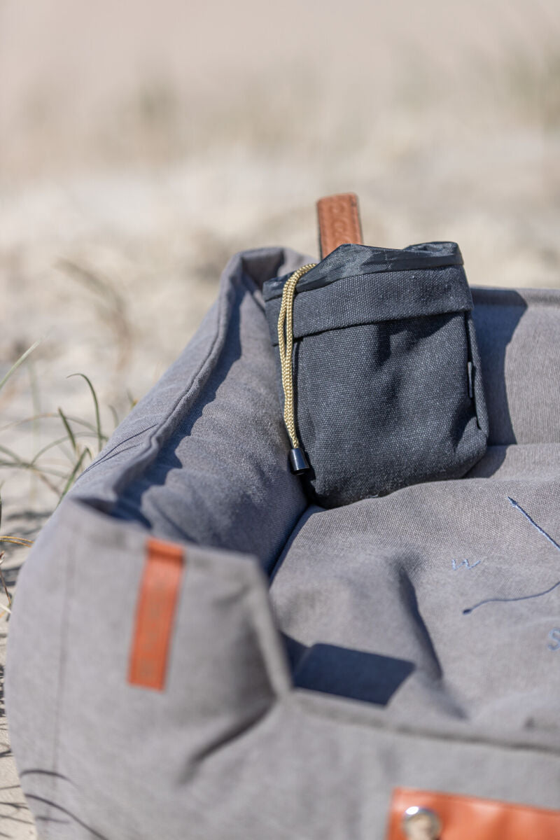 Graues Hundebett mit einer schwarzen Tasche auf sandigem Untergrund.