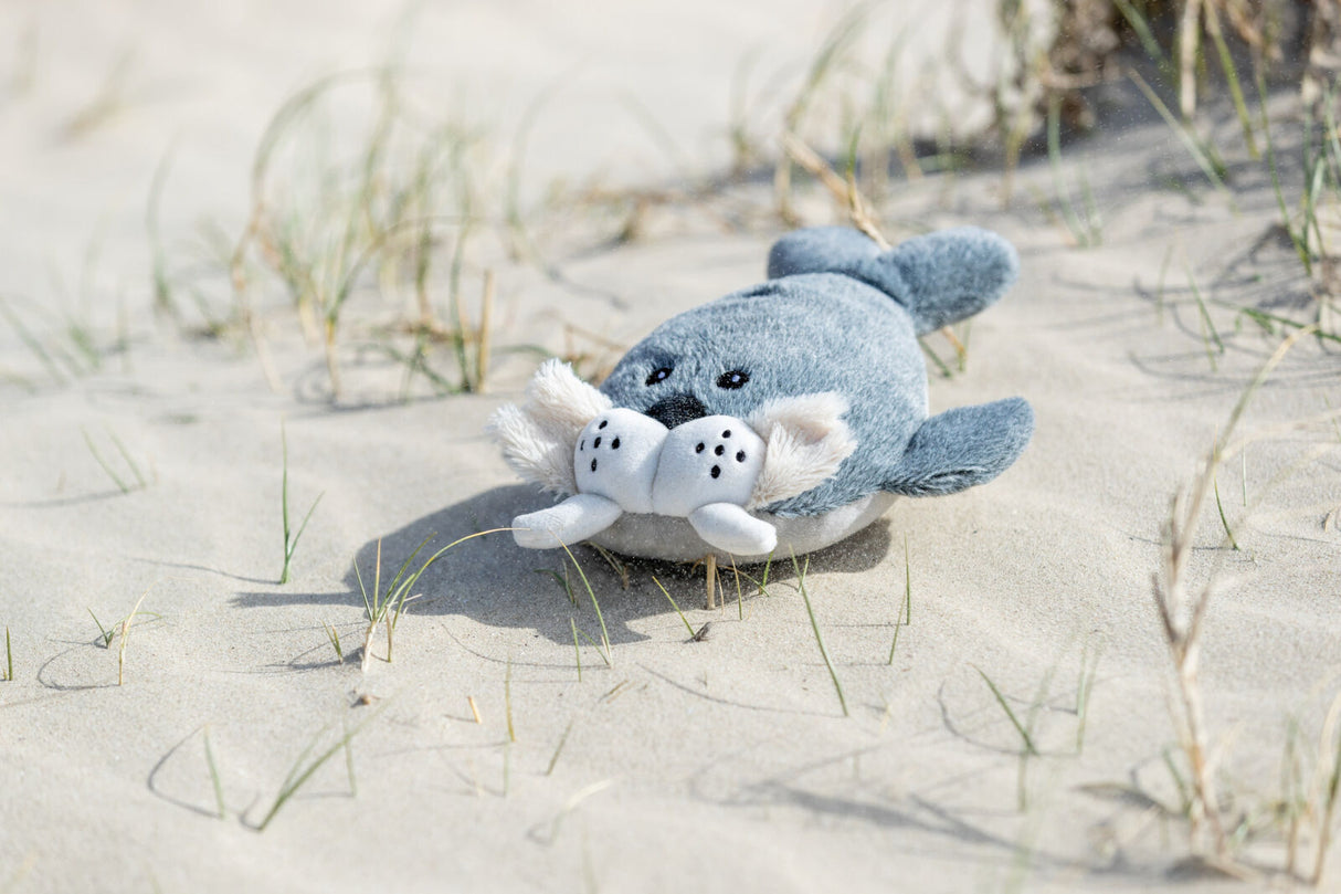 Ein graues Plüschtier in Form einer Robbe liegt auf Sand mit spärlichem Gras.