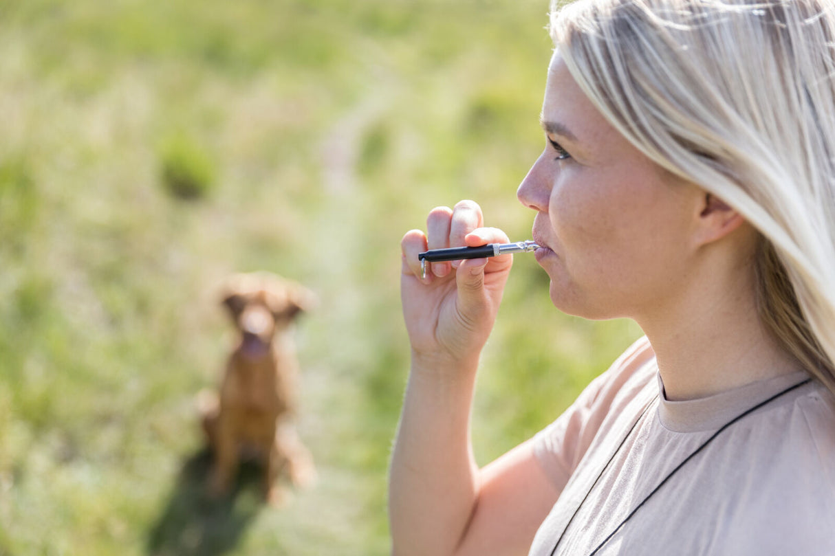 Eine Frau steht im Freien und bläst in eine Hundepfeife, im Hintergrund sitzt ein Hund.