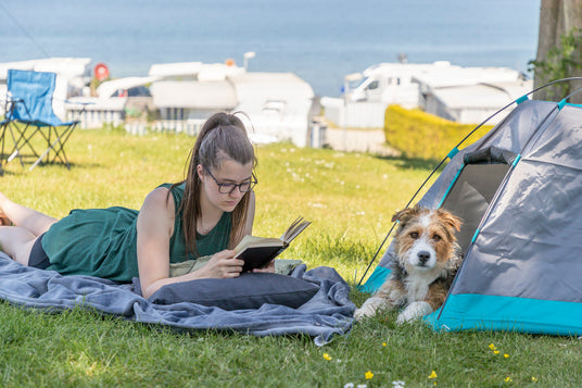Frau liegt auf dem Bauch auf einer Decke im Gras und liest ein Buch. Rechts daneben ist ein Hund in einem kleinen Hundezelt, schaut in die Kamera. Im Hintergrund scheint die Sonne und es ist der Campingplatz und das Meer zu sehen.