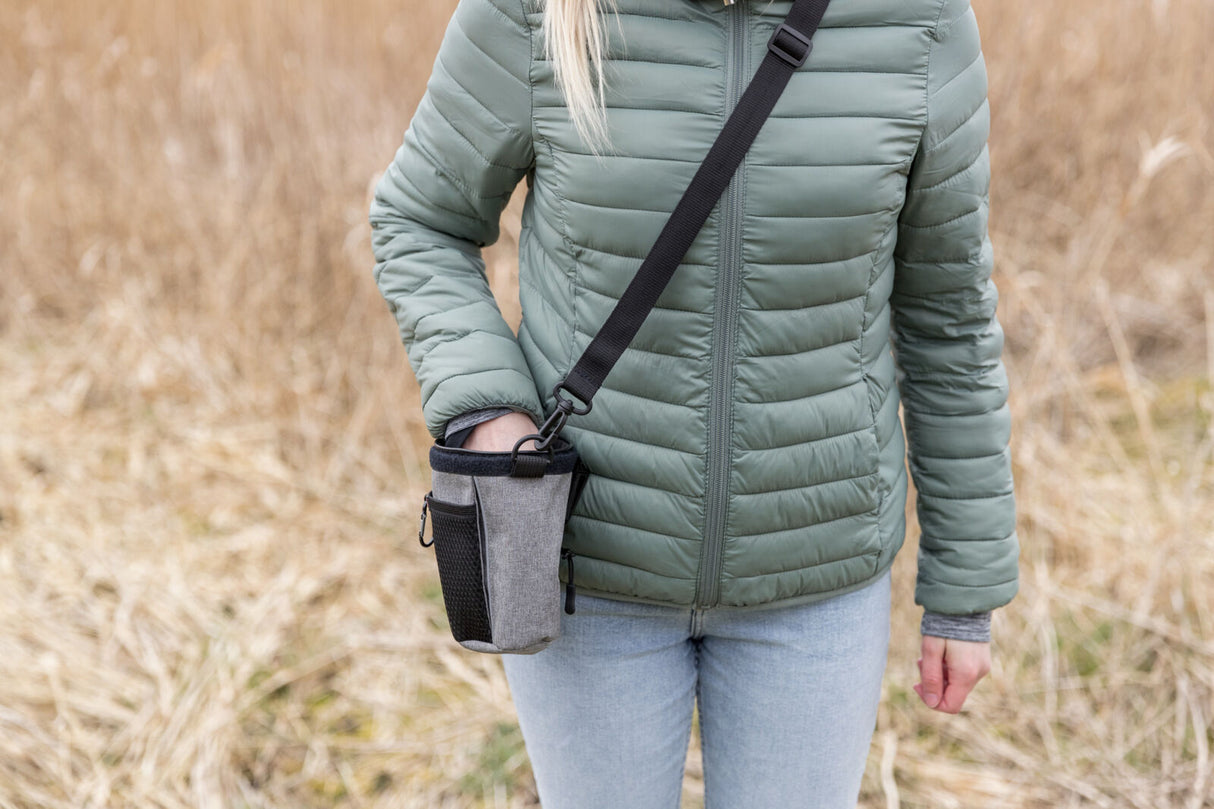 Person in grüner Jacke mit grauem Leckerlibeutel auf Feldweg im Freien.