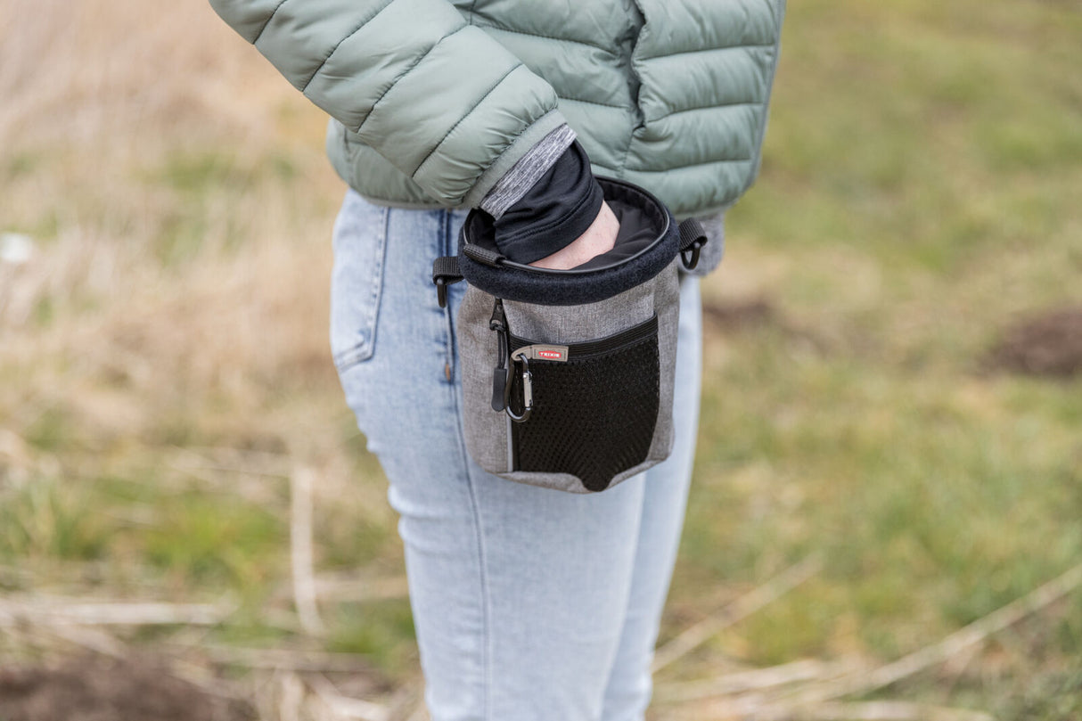 Person greift in eine kleine, graue Tasche mit schwarzem Netz auf einer Wiese.