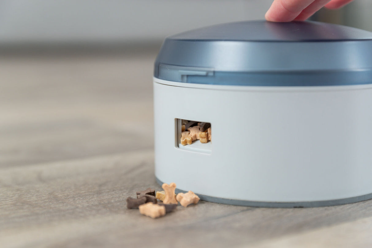 Ein weißer und grauer Behälter auf Tisch, teils offene Klappe, sichtbar Hundefutter, Hand auf Deckel.