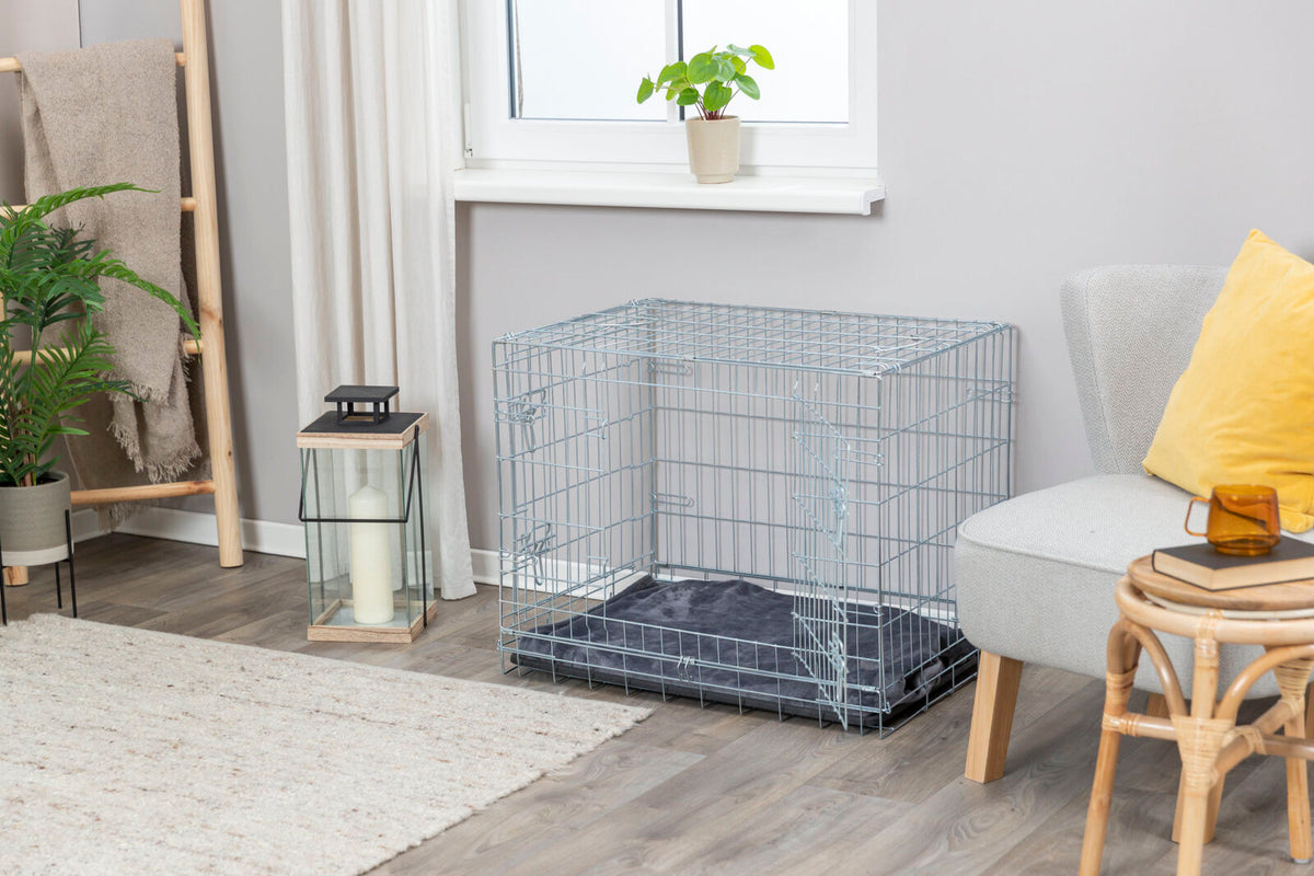 Metallene Hundebox mit offenem Gitterdesign steht auf Holzboden neben grauem Stuhl und gelbem Kissen.
