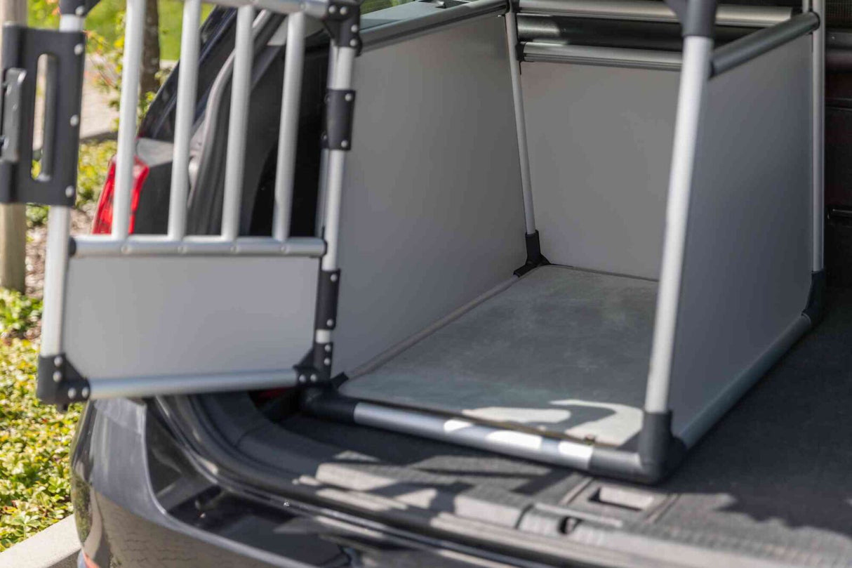 Geöffnete silberfarbene Transportbox mit Gittertür im Kofferraum eines Autos bei Tageslicht.