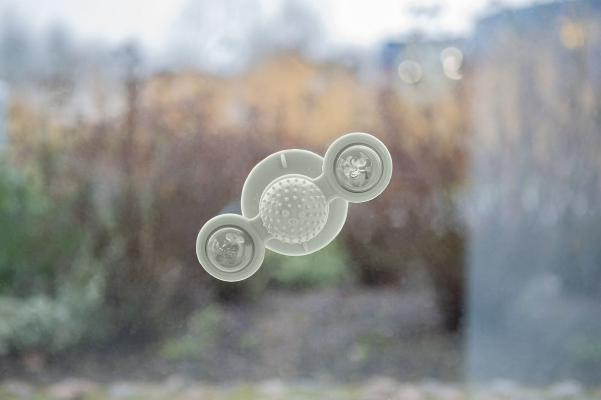 Ein rundes, weißes Spielzeug mit Saugnäpfen hängt an einer Fensterscheibe, draußen unscharfe Natur.