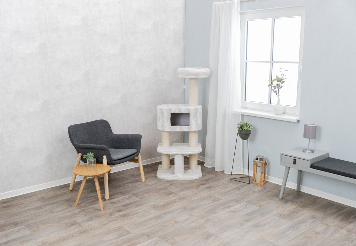 Wohnzimmer mit grauem Sessel, Holztisch, Kratzbaum und Fenster mit weißem Vorhang, Pflanzen auf Boden und Tisch.