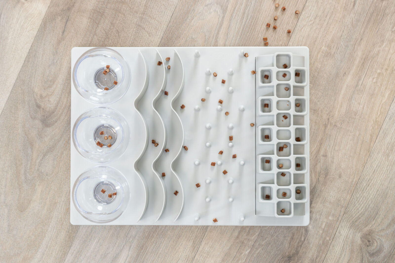 Weiße, wellenförmige Futterstation mit transparenten Schalen, verwendet für kleine braune Pellets auf hellem Holzboden.