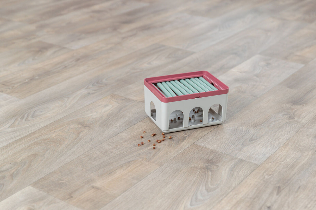 Ein weißer Behälter mit rotem Rand und grünlichen Rollen, auf Holzboden stehend, daneben verstreute Krümel.