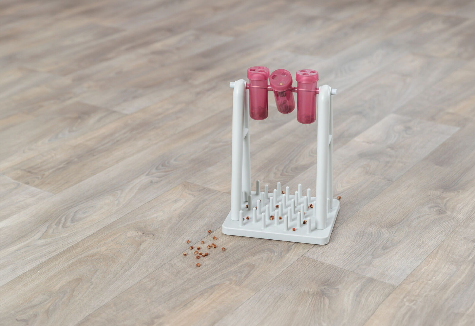 Weißer Ständer mit drei rosa Behältern und Stäben auf Holzboden, daneben verstreute braune Körner.