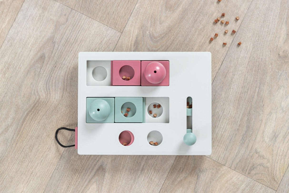 Weiße rechteckige Spielzeugbox mit farbigen Elementen und kleinen Leckerbissen auf einem Holzboden.