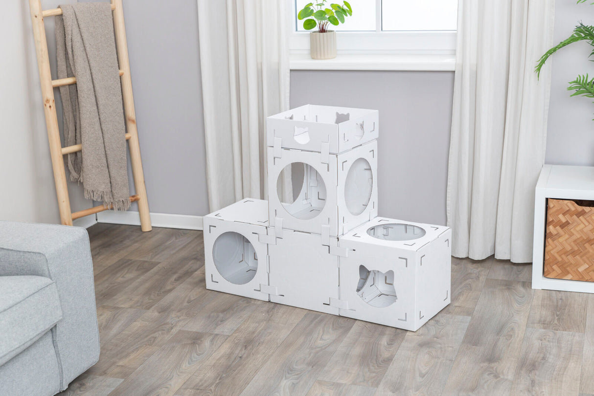 Weiße modulare Würfel mit kreisförmigen Öffnungen stehen in einem Zimmer auf einem Holzfußboden, im Hintergrund Pflanzen und ein graues Sofa.