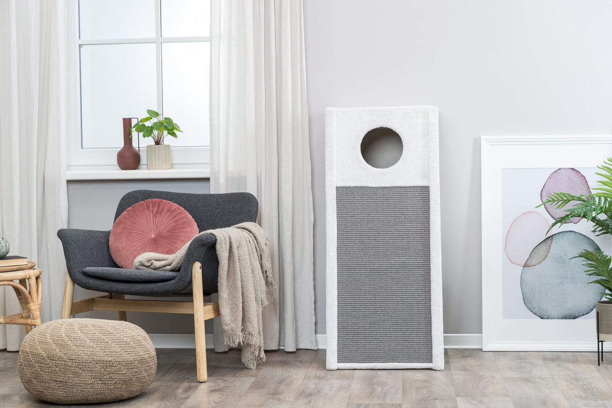 Ein grau-weißer Kratzbaum steht in einem hellen Raum neben einem grauen Sessel mit Kissen und Decke.