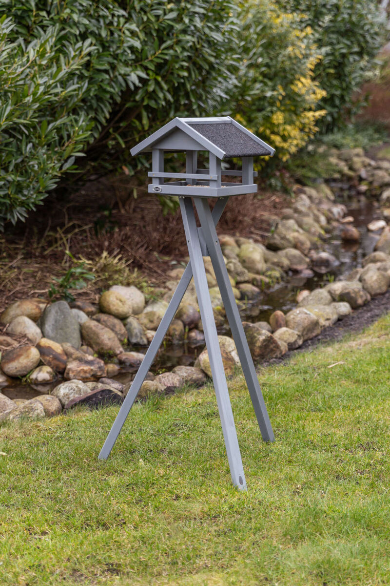 Graues Vogelhaus auf dreibeinigem Ständer im Garten, umgeben von Gras und Steinen, mit Büschen im Hintergrund.