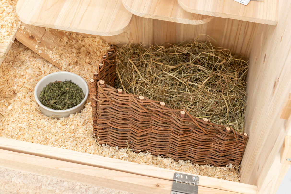 Ein Holzgehege mit Holzspänen, einem weißen Napf mit getrockneten Kräutern und einer geflochtenen Heuraufe.