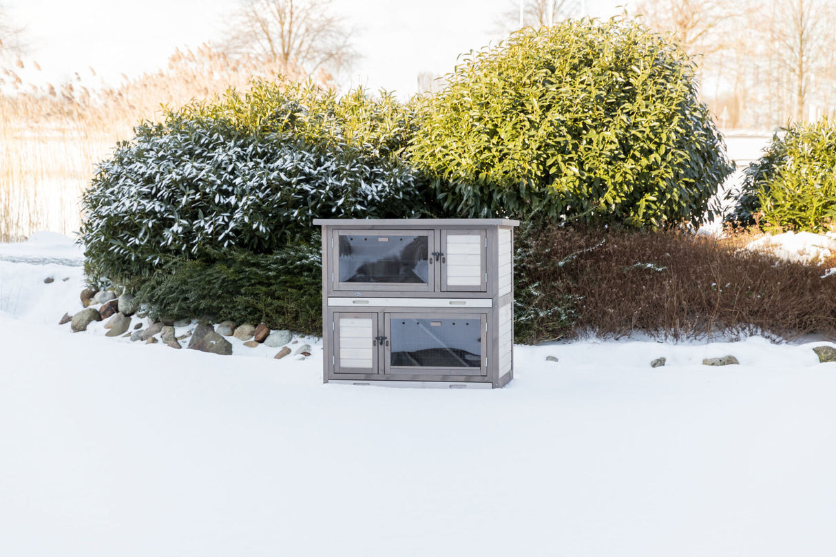 Ein graues, zweistöckiges Kleintiergehege steht im verschneiten Garten vor einem großen, grünen Strauch.