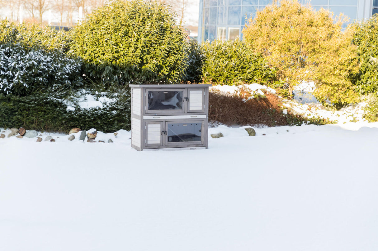 Grau-weißes zweistöckiges Holzgehege auf schneebedecktem Boden vor grünen Büschen im Freien.
