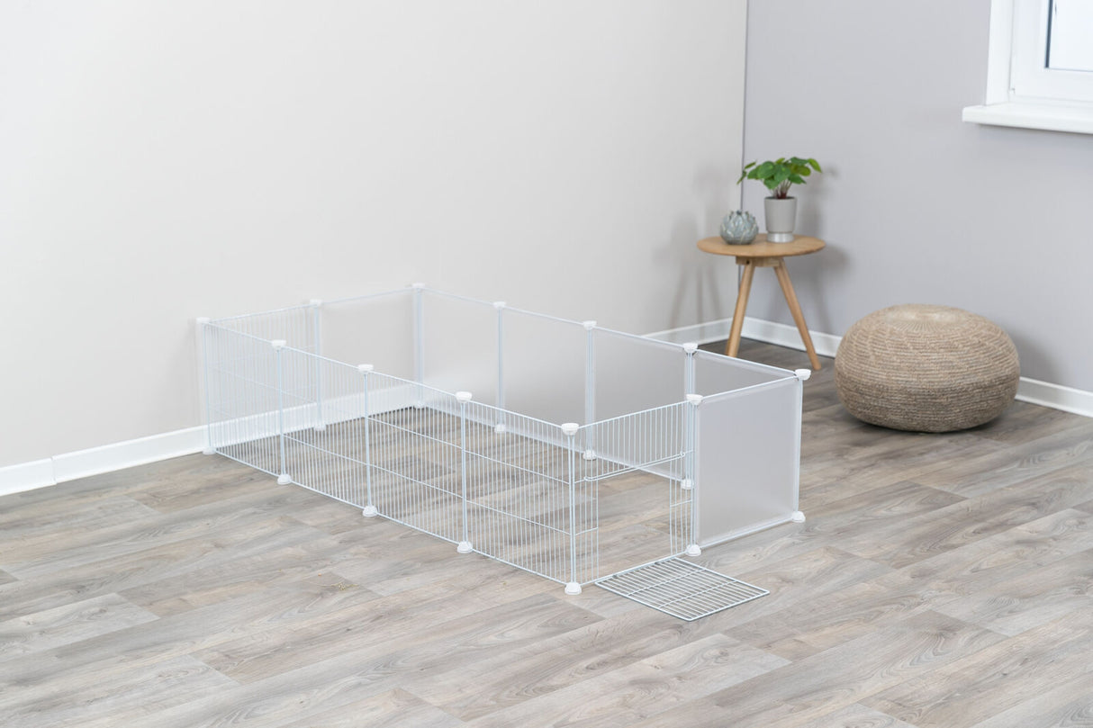 Ein weißer Gitterlaufstall steht auf einem Holzboden neben einem Tisch mit Pflanzen und einem runden Flechtkissen.