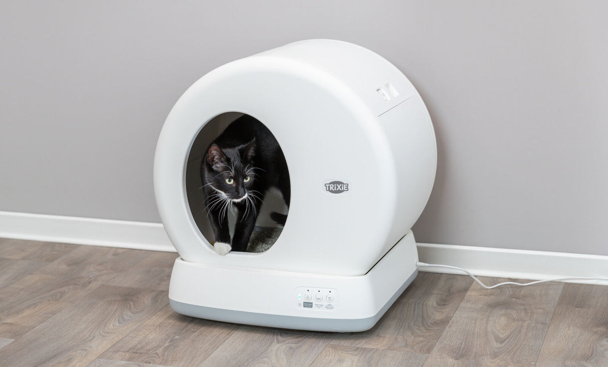 Schwarze Katze in einer runden, weißen Katzentoilette mit Trixie-Logo auf Holzfußboden; elektronisch verbunden.
