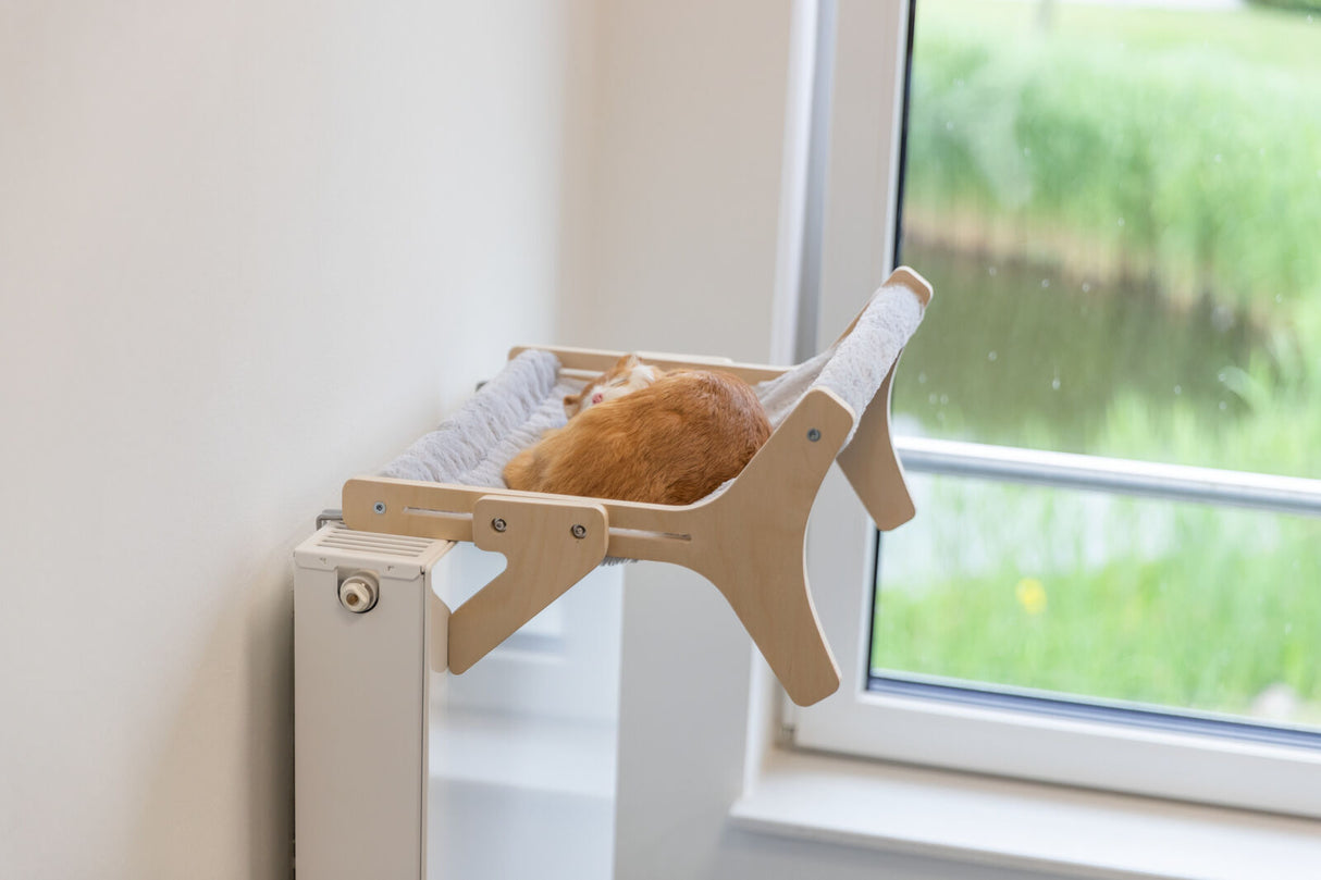 Eine Katze schläft auf einem an der Heizung befestigten Hängemattengestell neben einem Fenster.