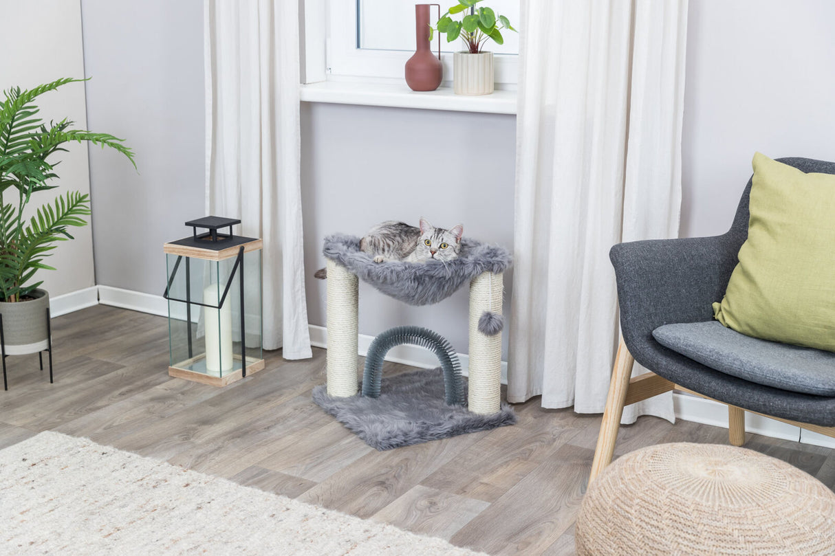 Eine graue Katze liegt auf einem kleinen, flauschigen Kratzbaum in einem hellen Raum mit Pflanzen und einem Sessel.