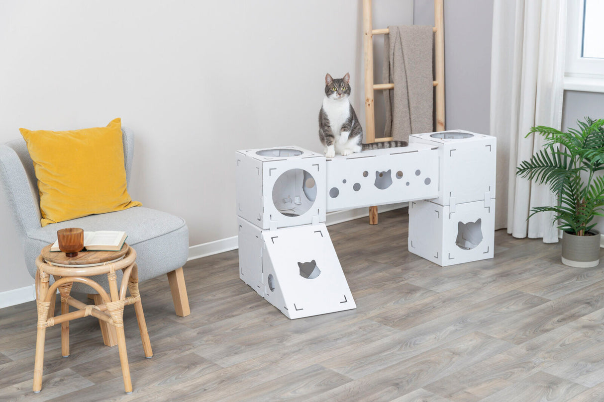 Eine grau-weiße Katze sitzt auf einem weißen modularen Katzenspielzeug neben einem Stuhl mit gelbem Kissen.