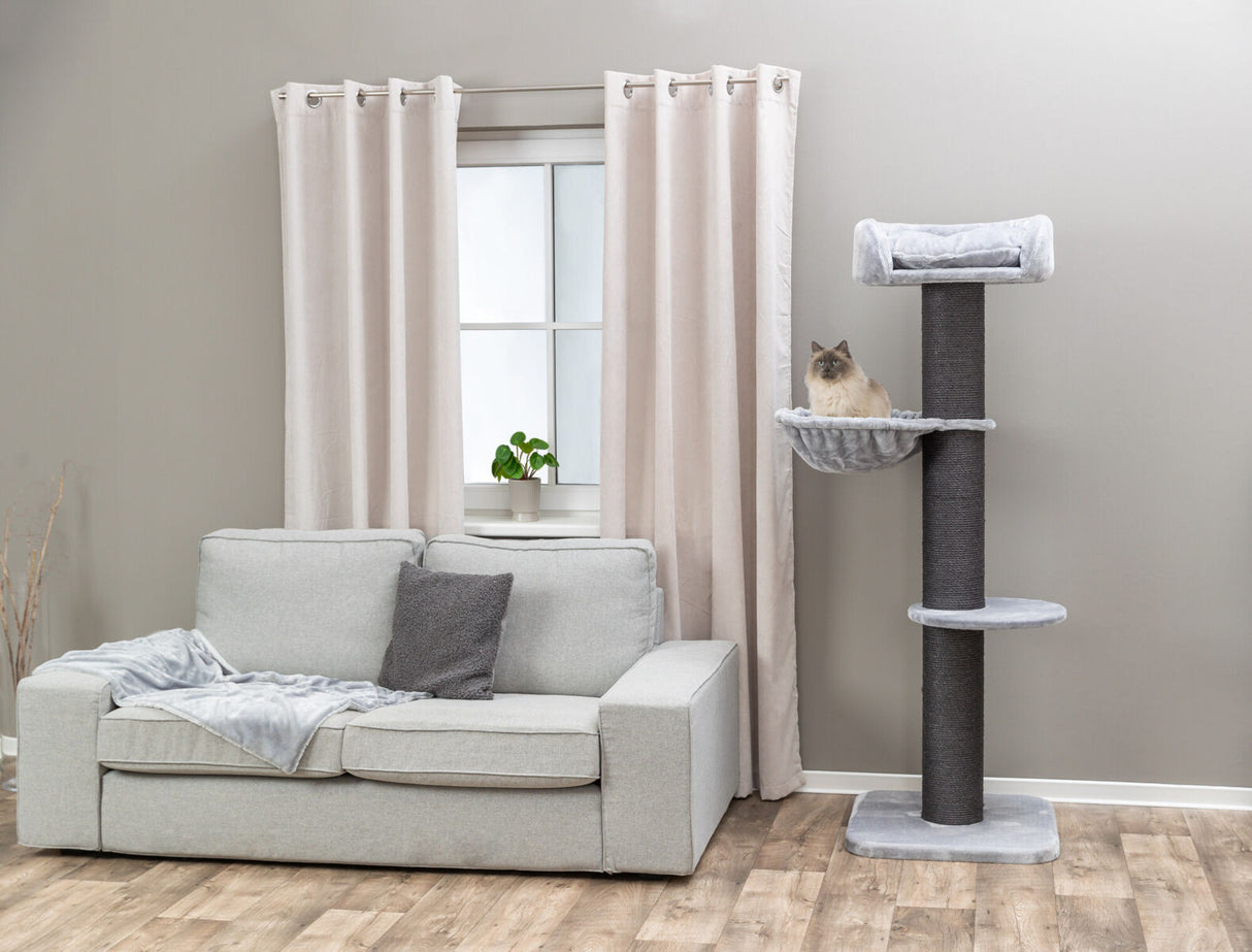Grauer Kratzbaum neben beiger Couch vor Vorhang und Fenster, Katze sitzt in erhöhter Liegefläche, Holzfußboden.