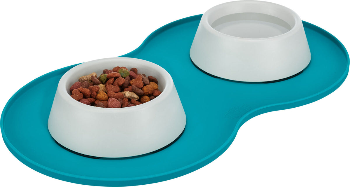 Zwei weiße Futternäpfe auf einer türkisfarbenen Matte, einer gefüllt mit Trockenfutter.