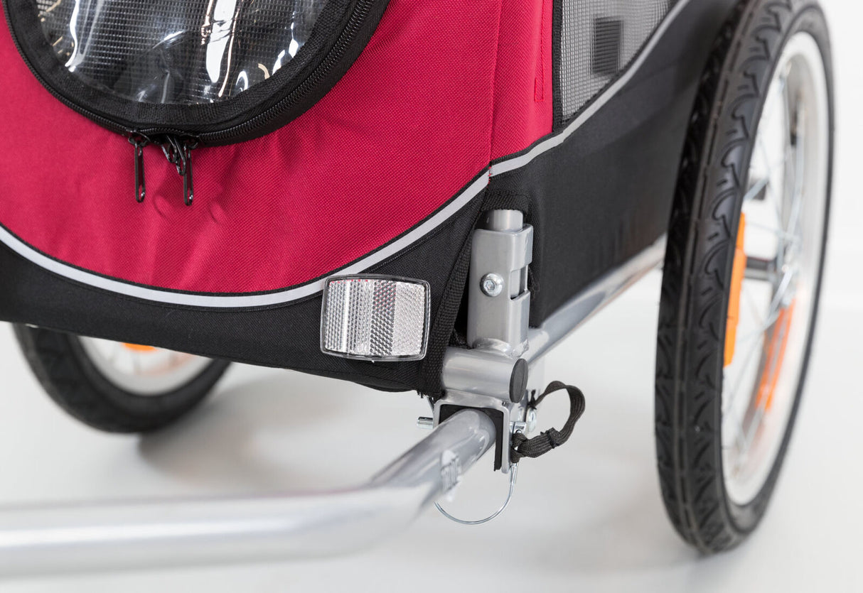 Ein rot-schwarzer Fahrradanhänger mit silbernem Rahmen, Reflektor und schwarzem Reifen auf weißem Hintergrund.