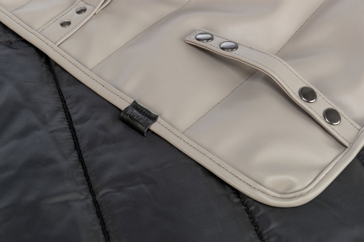 Beige Tasche mit silbernen Druckknöpfen auf schwarzem Hintergrund, Namenslabel aus Leder sichtbar.