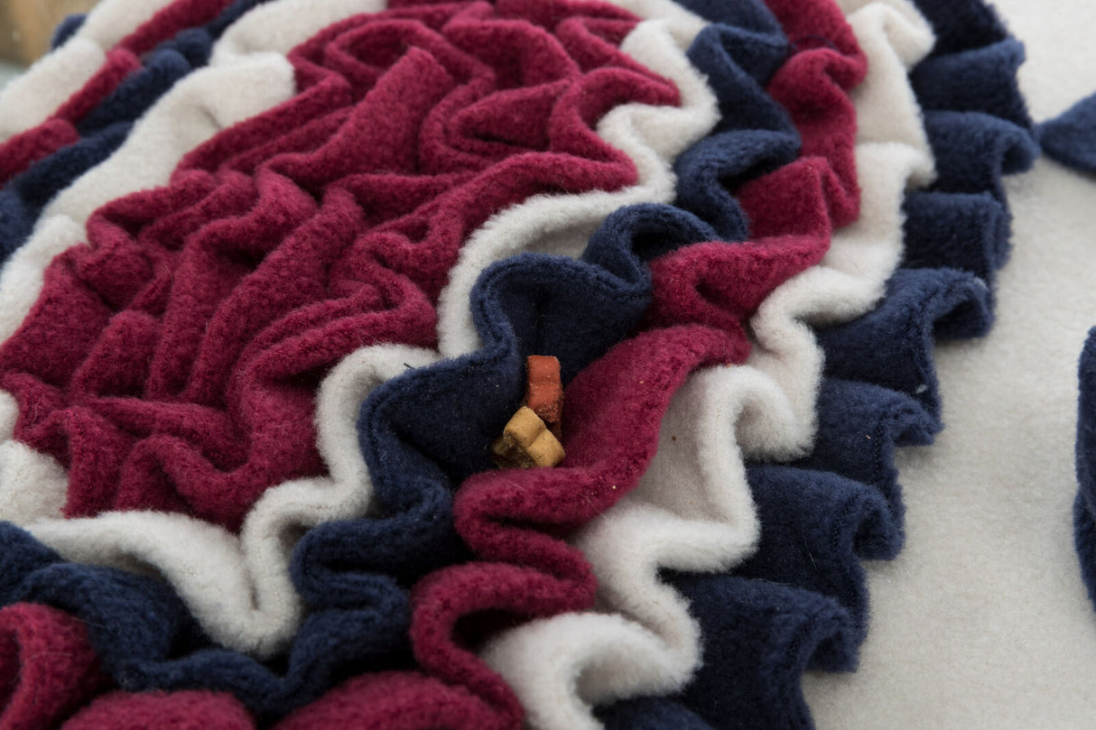 Bunte, ineinander verschlungene Filzschichten in Rot, Weiß und Blau mit kleinen Leckerli-Stücken in der Mitte.