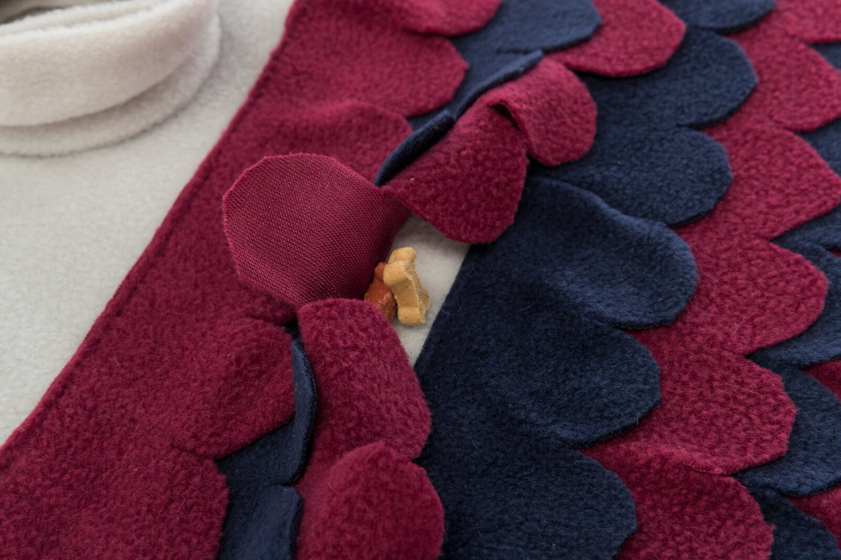 Roter und dunkelblauer Stoff mit wellenförmigen Kanten bedeckt einen cremefarbenen Hintergrund, darauf liegt ein kleiner, fleischfarbener Hundekeks.