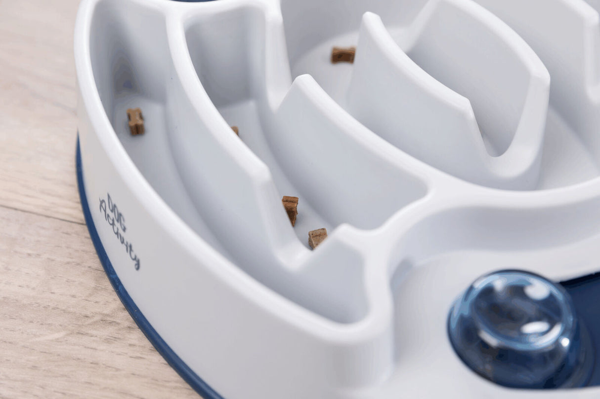 Weißer Futternapf in Labyrinthform mit braunen Stücken im Inneren auf einer Holzoberfläche.