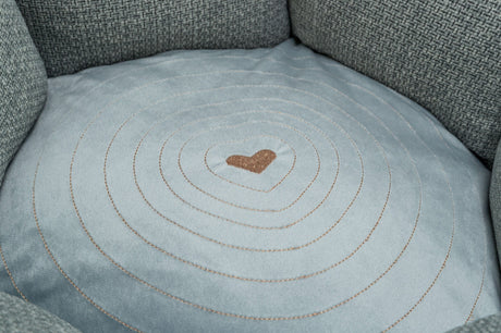 Graues, rundes Kissen mit hellbraunem, gesticktem Herz in der Mitte und konzentrischen Kreisen darauf.