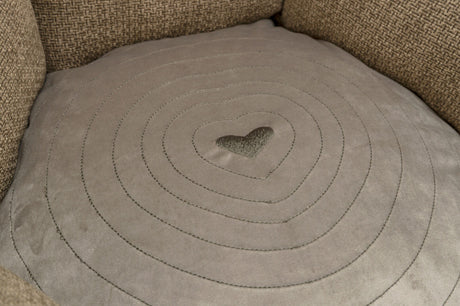 Graues Kissen mit Herzstickerei in der Mitte und konzentrischen Kreisen, umgeben von grobem, braunem Stoff.