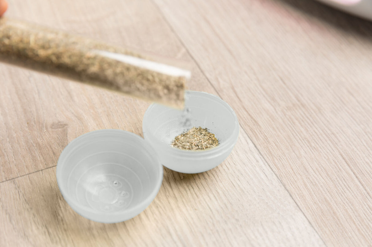 Kräuter werden aus einem Reagenzglas in eine geöffnete graue Plastikschale auf einem Holztisch geschüttet.