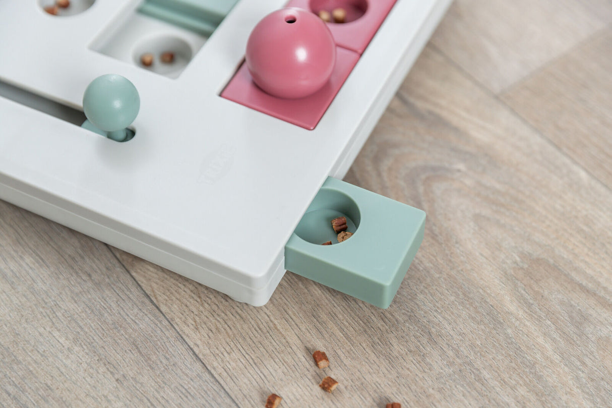 Ein weißes Puzzle-Spielzeug mit roten und blauen Elementen und Fächern, teilweise mit kleinen braunen Snacks gefüllt.