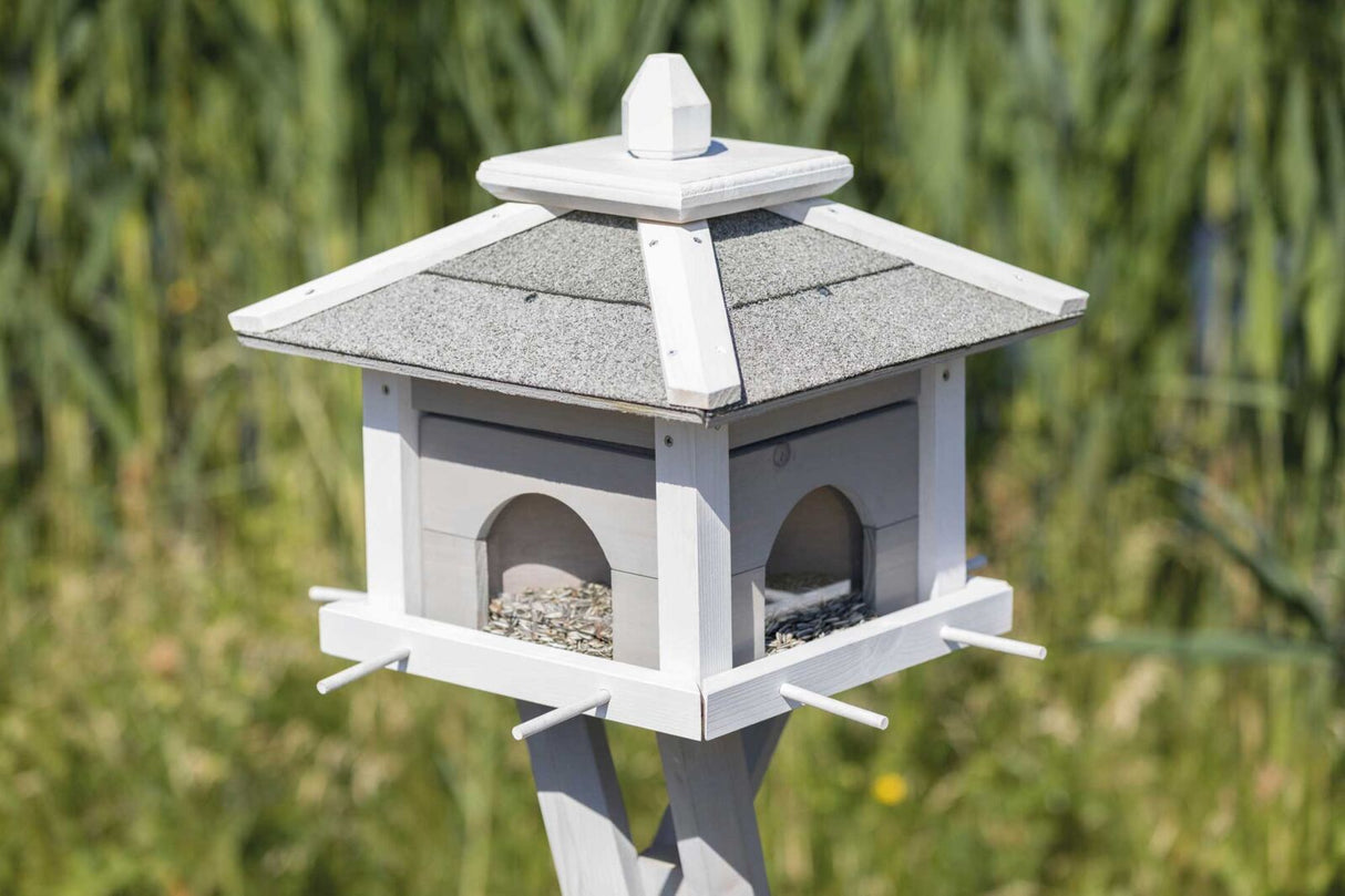 Ein graues und weißes Vogelhaus aus Holz mit schwarz-grauem Dach steht auf einem Holzpfosten im Freien.