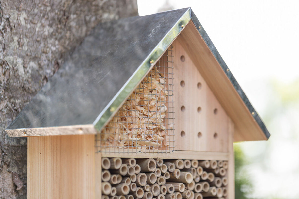 Holzinsektenhotel mit Metallrahmen und Holzröhren unter einem schrägen Blechdach an einem Baum.
