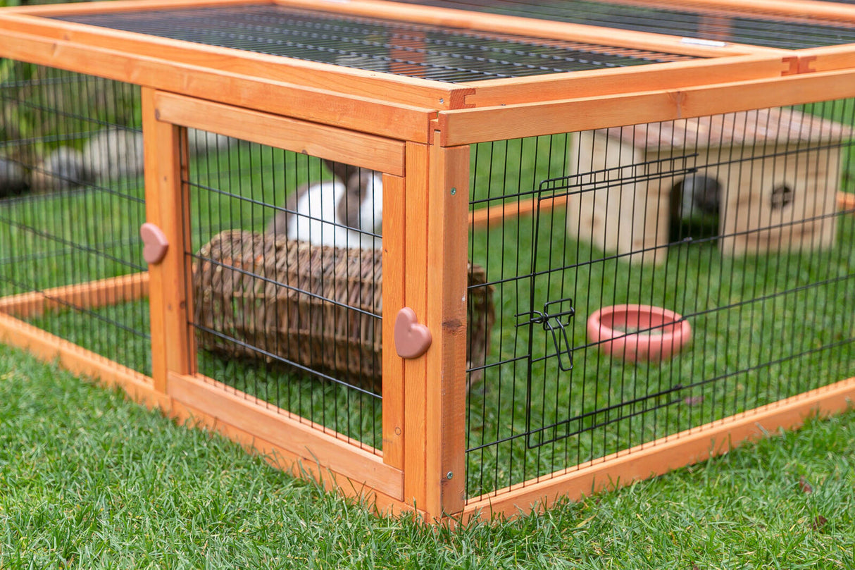 Holzgehege auf Gras mit Gitterwänden, darin ein Kaninchen, ein geflochtener Tunnel, ein Häuschen und ein Futternapf.