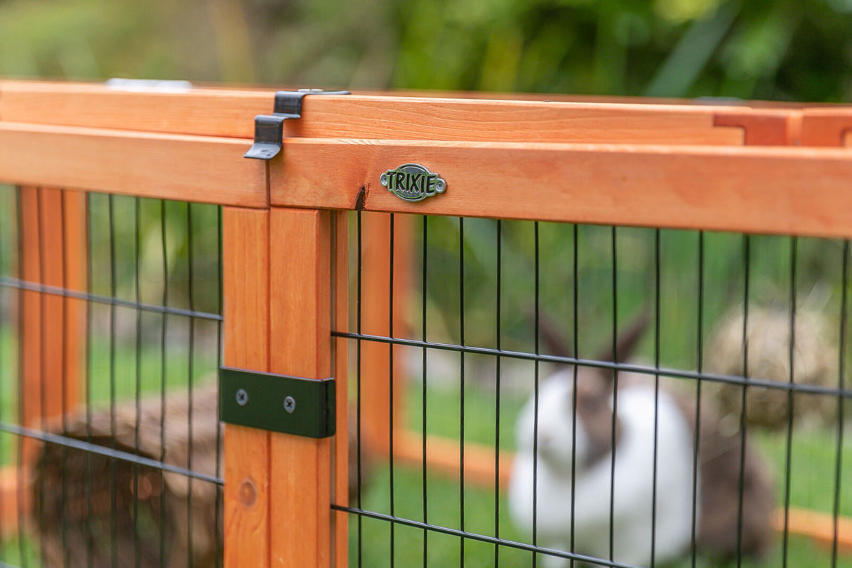 Holzgehege mit Gitterstäben und Trixie-Logo, im Hintergrund unscharfer weiß-brauner Hase auf Gras.