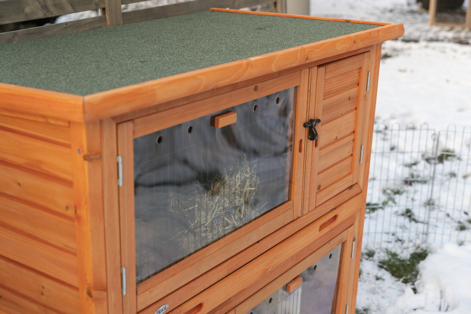 Holzstall mit grünem Dach, transparenten Klappen und schwarzem Verschluss, steht im schneebedeckten Außenbereich.