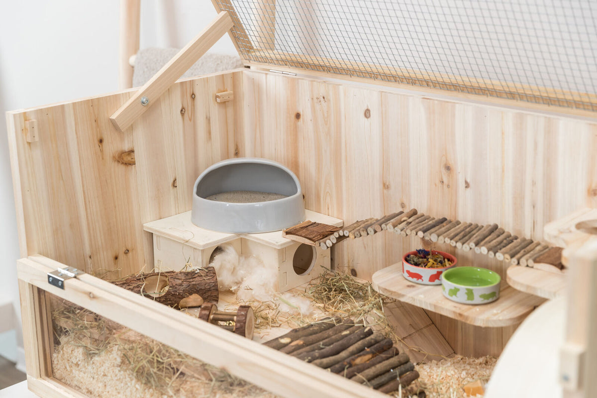 Holzgehege mit Streu und Heu, Brücke aus Zweigen, grauer Napf, Spielzeug, zwei bunte Futterschalen auf Plattform.