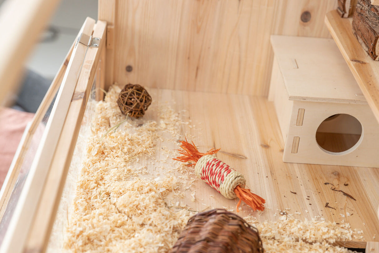 Kleiner Holzkäfig mit Streu, Spielzeug, Weidenkugel und Holzhaus, alles auf Holzfläche sichtbar.