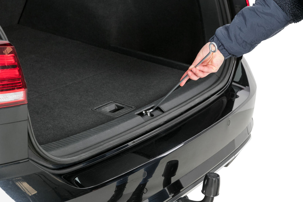 Eine Hand hebt mit einem Ringwerkzeug eine Abdeckung im Kofferraum eines schwarzen Autos an.