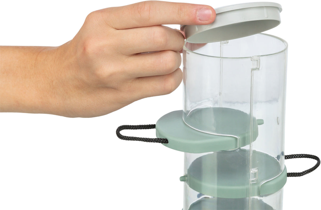 Hand öffnet einen transparenten, zylindrischen Behälter mit grünem Deckel und mehreren Ebenen, schwarzer Kordelgriff.