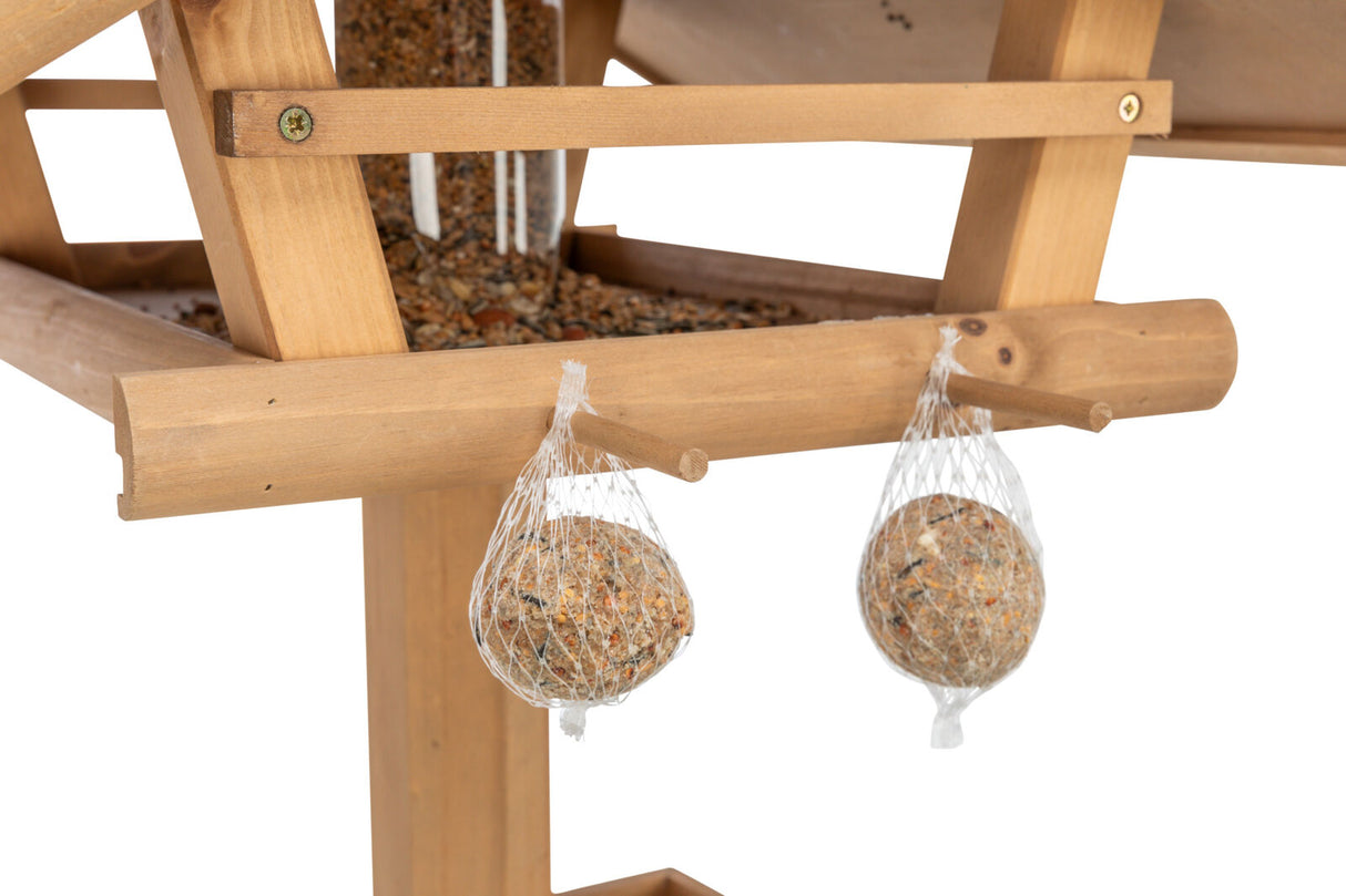 Nahaufnahme eines hölzernen Vogelhäuschens mit Vogelfutter und zwei Meisenknödeln in Netzbeuteln darunter.
