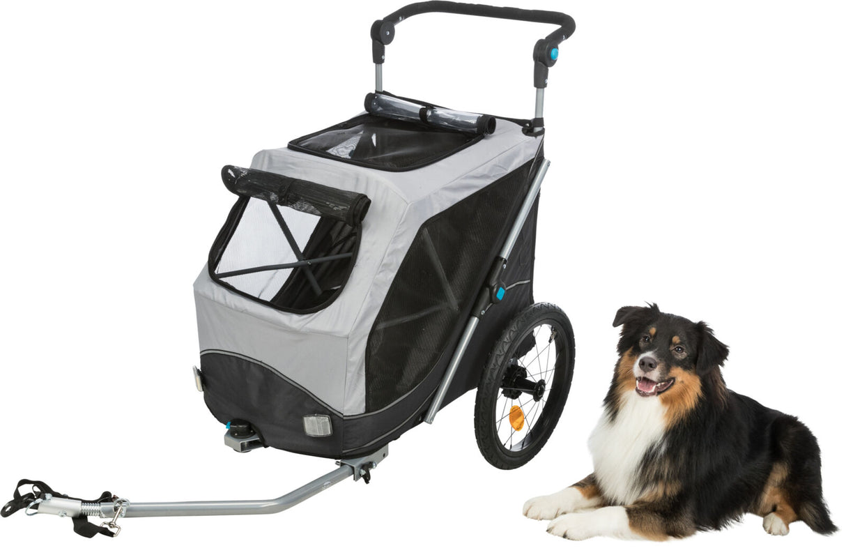 Grauer Fahrradanhänger mit schwarzen Rädern neben einem sitzenden dreifarbigen Hund auf weißem Hintergrund.
