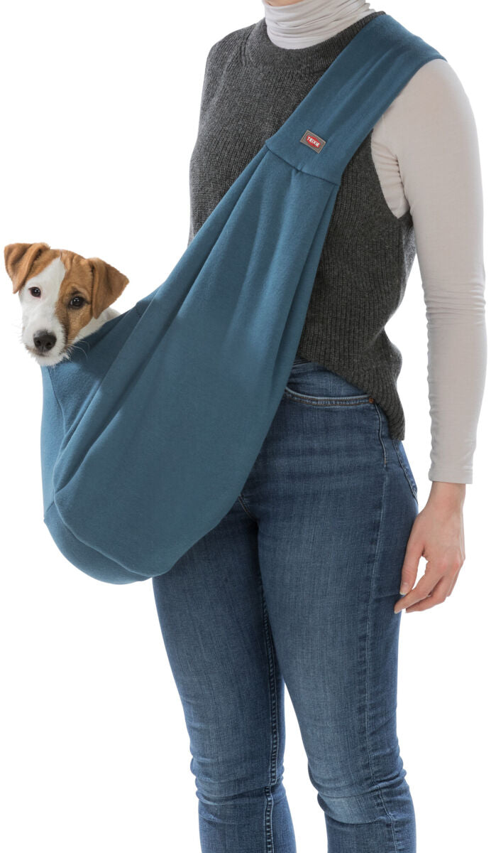 Person trägt eine blaue Tragetasche mit einem Hund darin, während sie eine graue Weste und blaue Jeans trägt.